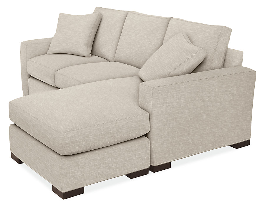 The Metro Sofas with Chaise is a light beige sectional featuring two seat cushions and a left-side chaise lounge. It comes with three back cushions and two matching throw pillows, all supported by dark wood legs and boasting a subtle woven fabric texture.