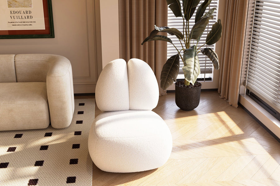 A modern living room features the Moony Fleece Curved Lounge Chair by Acanva on a light wood floor. A beige couch is partially visible on the left, positioned next to a framed artwork. A large potted plant stands near the window, adorned with beige curtains and blinds that allow sunlight to filter in.
