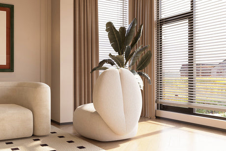 A modern living room showcases the Moony Fleece Curved Lounge Chair by Acanva, designed with a striking cushion arrangement resembling abstract petals, complemented by a potted plant rising from the center. The space is adorned with beige curtains and features a large window equipped with blinds, along with a cream-colored sofa.