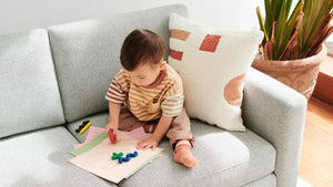 A toddler sits on a Nomad Fabric Sofa, drawing on papers with colorful markers. A white pillow with geometric patterns is beside him, and a plant is in the background. The scene is lit by natural sunlight from a nearby window.