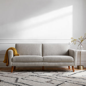 In a contemporary living room, the Oliver Sofa in light gray with wooden legs sits against a white wall. A mustard yellow blanket is elegantly draped over its armrest. Beside the sofa, there's a round side table adorned with a vase of branches. A patterned area rug decorates the floor.