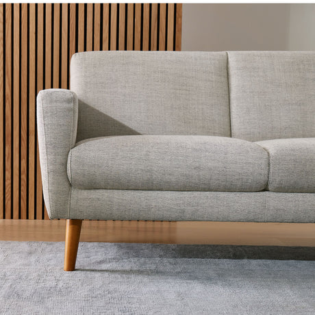 The Oliver Sofa, featuring light gray upholstery and wooden legs, is displayed in a living room. The backdrop includes a wooden slat wall, while a light gray carpet covers the floor, showcasing a minimalist, contemporary design style.