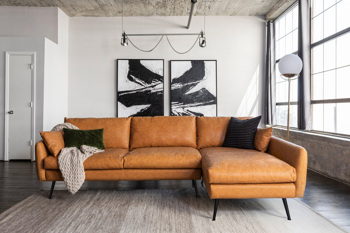 A modern living room showcases the Park Sectional Sofa in a spacious tan leather design, accented with a black and green pillow and complemented by a draped beige throw. Large abstract black-and-white paintings adorn the wall, while sunlight pours through tall windows.