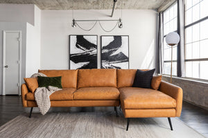 A modern living room showcases the Park Sectional Sofa in a spacious tan leather design, accented with a black and green pillow and complemented by a draped beige throw. Large abstract black-and-white paintings adorn the wall, while sunlight pours through tall windows.