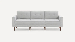 The Nomad Fabric Sofa is a contemporary gray piece with a minimalist design, featuring three cushions and elegant wooden legs, set against a simple backdrop.