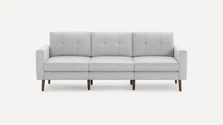 The Nomad Fabric Sofa is a contemporary gray piece with a minimalist design, featuring three cushions and elegant wooden legs, set against a simple backdrop.