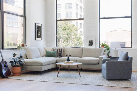 A bright, modern living room featuring a spacious Sloan Custom 3-Seat 95" cream sofa in an L-shape, alongside a gray armchair and a round wooden coffee table. The space is complemented by lush plants and a guitar, with large windows allowing natural light to flood in. The minimalist decor is accented by framed art on the walls.