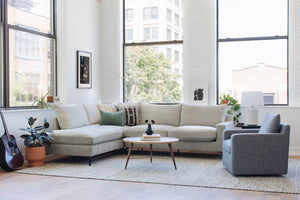 A bright, modern living room featuring a spacious Sloan Custom 3-Seat 95" cream sofa in an L-shape, alongside a gray armchair and a round wooden coffee table. The space is complemented by lush plants and a guitar, with large windows allowing natural light to flood in. The minimalist decor is accented by framed art on the walls.