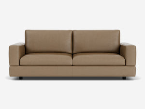 The Stage Track Sofa, featuring a minimalist design with thick cushions and square armrests, is showcased against a white background. Its brown leather upholstery and sleek, contemporary style are enhanced by short black legs.