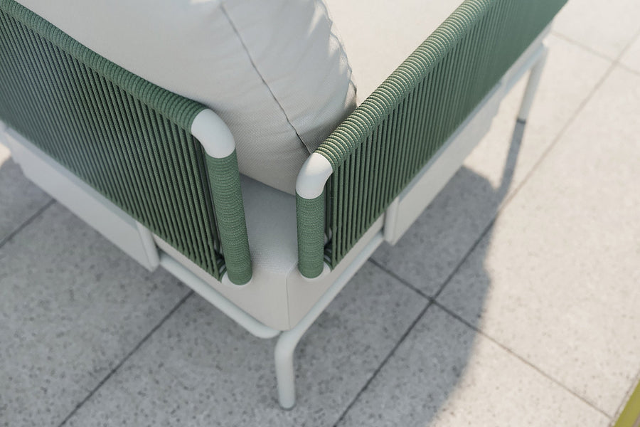 Close-up of the Sylva Outdoor Modular Sofa by Acanva, featuring green woven rope detailing and white cushions. The sofa rests on a light stone tile floor, casting a shadow in the sunlight.