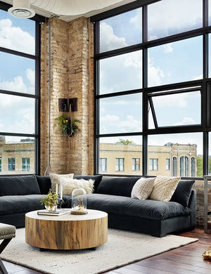 A modern living room with large windows showcasing a cityscape. It features the Decima Sectional Sofa, a dark L-shaped piece adorned with cushions, along with a round wooden coffee table decorated tastefully, and brick walls that allow natural light to fill the space.